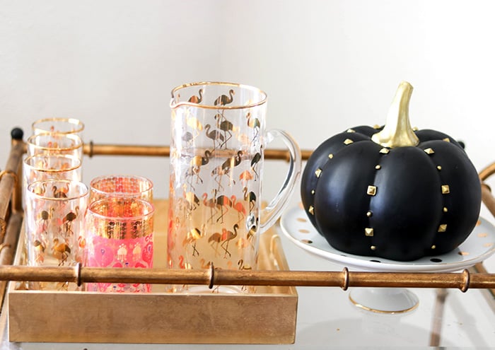 Candie Anderson decorated her fall bar cart with a black gold studded pumpkin, flamingo glassware and a white cake stand with gold polka dots.