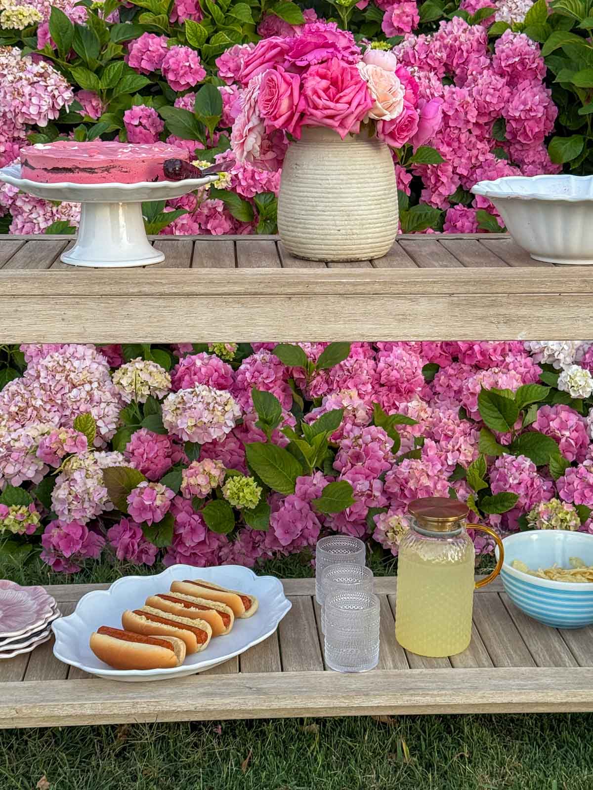 Entertaining expert, journalist and lifestyle blogger Candie Anderson of candieanderson.com shares her summer entertaining station with a white platter adorned with hotdogs, a pitcher filled with lemonade, a ceramic vase with hydrangeas and a red velvet ice cream cake on a white cake stand.