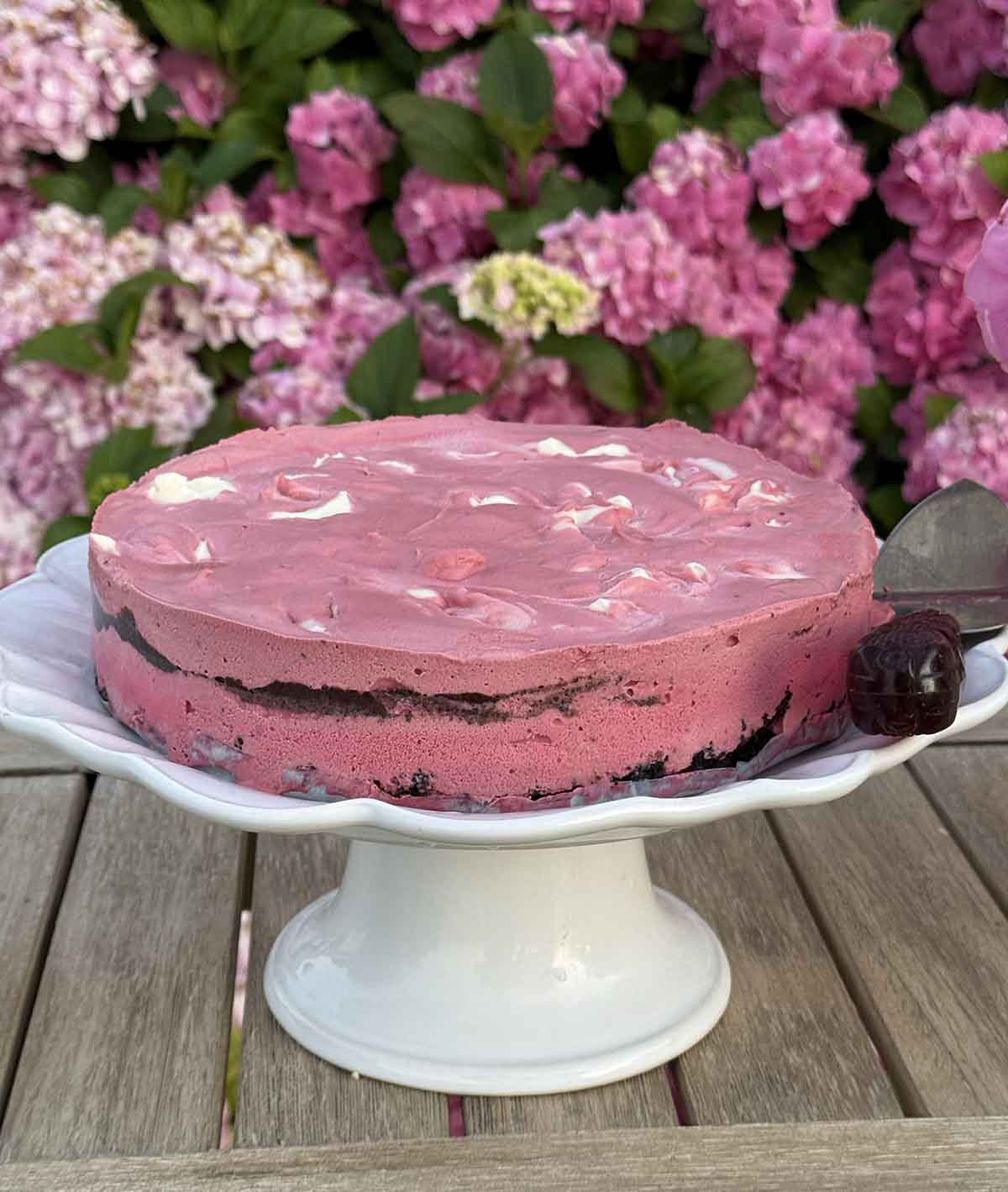 Entertaining expert, journalist and lifestyle blogger Candie Anderson of candieanderson.com shares the best homemade red velvet ice cream cake on a white cake stand in her hydrangea garden.